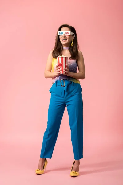 Full length view of happy young stylish disco girl in 3d glasses holding striped paper bucket with popcorn on pink — Stock Photo