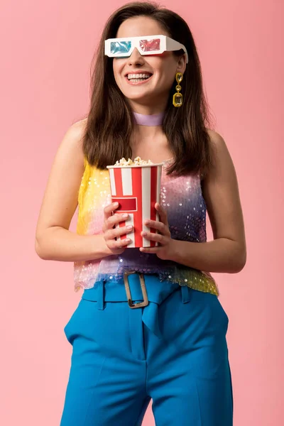Happy young stylish disco girl in 3d glasses holding striped paper bucket with popcorn isolated on pink — Stock Photo
