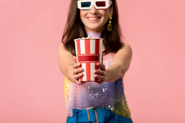 Enfoque selectivo de la joven chica disco con estilo feliz en gafas 3d presentando cubo de papel rayado con palomitas de maíz aislado en rosa - foto de stock