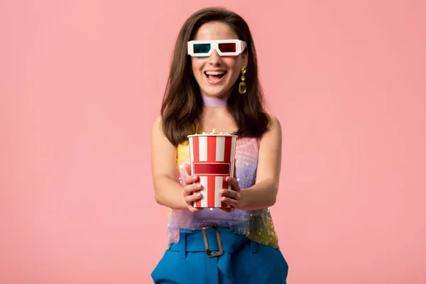 Happy young stylish disco girl in 3d glasses holding striped paper bucket with popcorn isolated on pink — Stock Photo