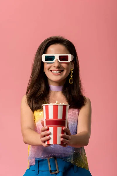 Happy young stylish disco girl in 3d glasses holding striped paper bucket with popcorn isolated on pink — Stock Photo