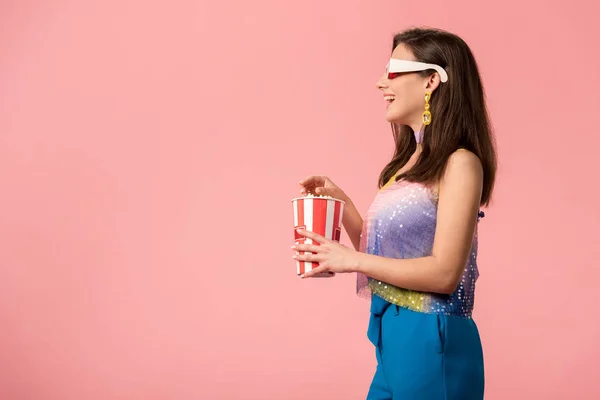 Вид збоку щасливої молодої стильної дівчини диско в 3d окулярах їсть попкорн ізольовано на рожевому — стокове фото