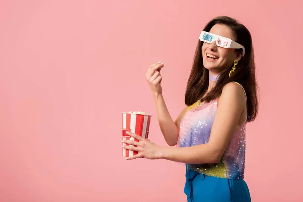 Vista lateral de la joven y feliz disco chica con estilo en gafas 3d comer palomitas de maíz aislado en rosa - foto de stock