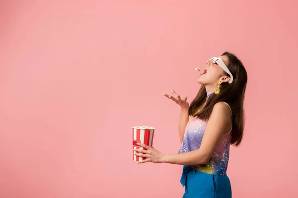 Vista lateral de la joven y feliz disco chica con estilo en gafas 3d lanzando palomitas de maíz en la boca aislado en rosa - foto de stock