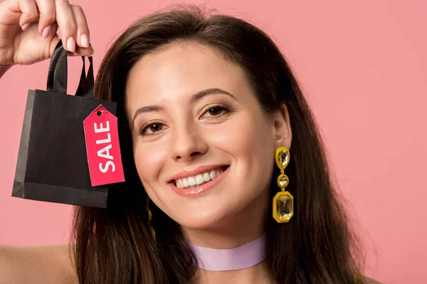 Happy stylish disco girl holding shopping bag with sale lettering on label isolated on pink — Stock Photo