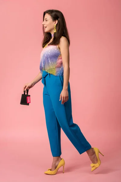 Full length view of stylish disco girl holding shopping bag with sale lettering on label on pink — Stock Photo