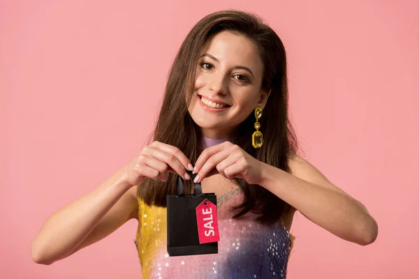 Happy stylish disco girl holding shopping bag with sale lettering on label isolated on pink — Stock Photo