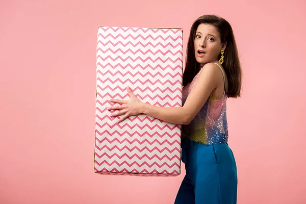 Schockiertes junges stilvolles Disco-Mädchen hält Geschenkbox isoliert auf rosa — Stockfoto