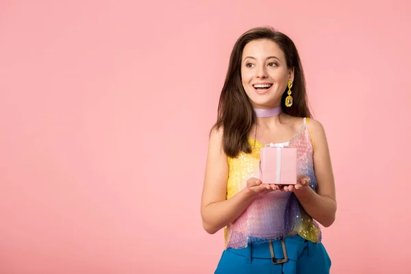 Feliz joven con estilo disco chica sosteniendo caja de regalo y mirando hacia otro lado aislado en rosa - foto de stock