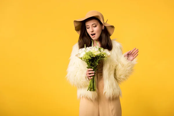 Shocked elegant girl in faux fur jacket and hat looking at bouquet isolated on yellow — Stock Photo
