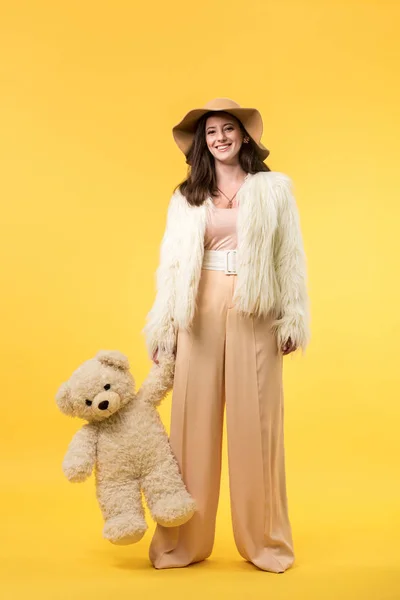 Heureuse fille élégante en fausse veste de fourrure et chapeau tenant ours en peluche sur jaune — Photo de stock