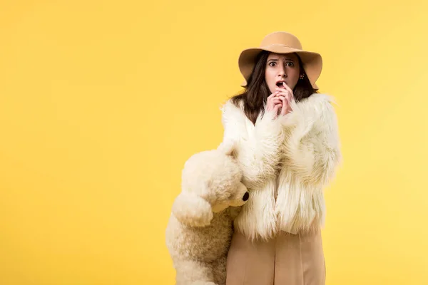 Shocked elegant girl in faux fur jacket and hat holding teddy bear isolated on yellow — Stock Photo