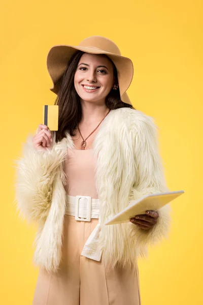 Happy elegant girl in faux fur jacket and hat holding digital tablet and credit card isolated on yellow — Stock Photo