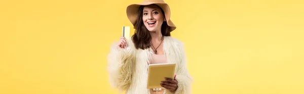 Панорамний знімок щасливої елегантної дівчини в хутряній куртці і капелюсі, що тримає цифровий планшет і кредитну картку ізольовано на жовтому — стокове фото