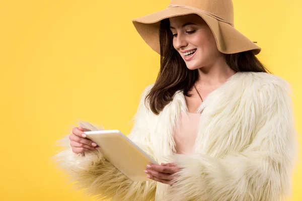 Chica elegante feliz en chaqueta de piel sintética y sombrero usando tableta digital aislado en amarillo - foto de stock
