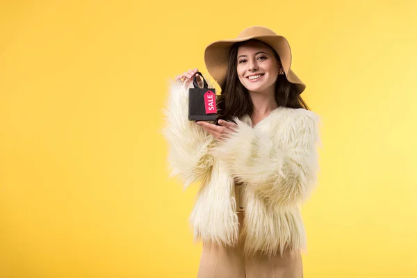 Chica elegante sonriente en chaqueta de piel sintética y sombrero sosteniendo bolsa de compras con letras venta aislado en amarillo - foto de stock