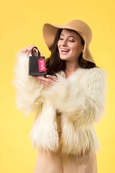Happy elegant girl in faux fur jacket and hat holding shopping bag with sale lettering isolated on yellow — Stock Photo