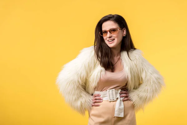 Smiling party girl in faux fur jacket and sunglasses with hands on hips isolated on yellow — Stock Photo