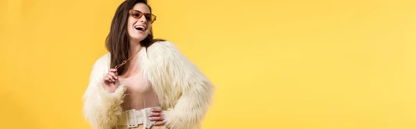 Panoramic shot of excited party girl in faux fur jacket and sunglasses isolated on yellow — Stock Photo