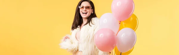 Colpo panoramico di festa felice ragazza in giacca di pelliccia sintetica e occhiali da sole tenendo palloncini isolati su giallo — Foto stock