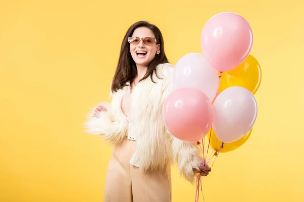 Felice festa ragazza in giacca di pelliccia sintetica e occhiali da sole in possesso di palloncini isolati su giallo — Foto stock