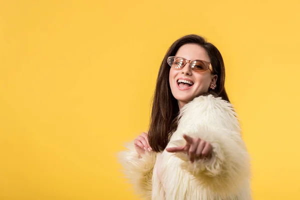 Ragazza partito eccitato in giacca di pelliccia sintetica e occhiali da sole che punta con il dito alla fotocamera isolata sul giallo — Foto stock