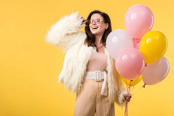 Счастливая девушка партии в искусственной меховой куртке и солнцезащитных очках держа воздушные шары изолированы на желтый — стоковое фото