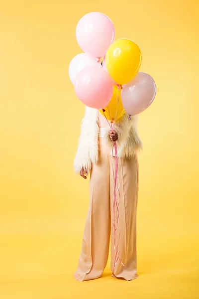 Chica de fiesta en chaqueta de piel sintética sosteniendo globos en frente de la cara sobre fondo amarillo - foto de stock