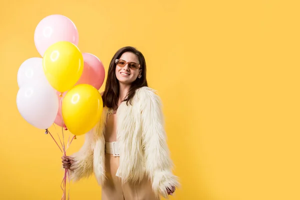 Chica de fiesta sonriente en chaqueta de piel sintética y gafas de sol sosteniendo globos aislados en amarillo - foto de stock