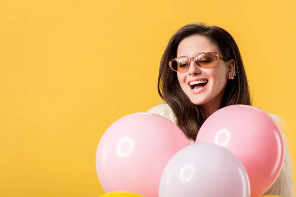 Felice festa ragazza in giacca di pelliccia sintetica e occhiali da sole in possesso di palloncini isolati su giallo — Foto stock