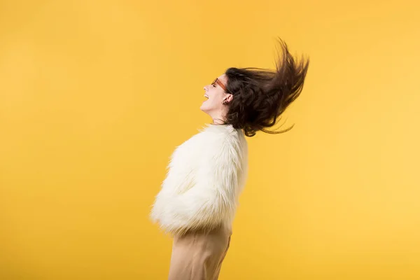 Side view of happy girl in faux fur jacket and sunglasses shaking head isolated on yellow — Stock Photo