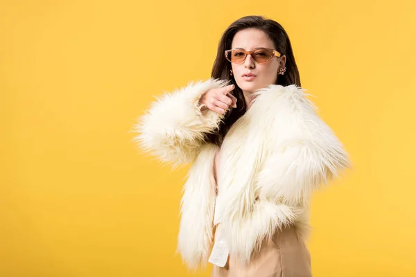 Girl in faux fur jacket and sunglasses pointing with finger at camera isolated on yellow — Stock Photo