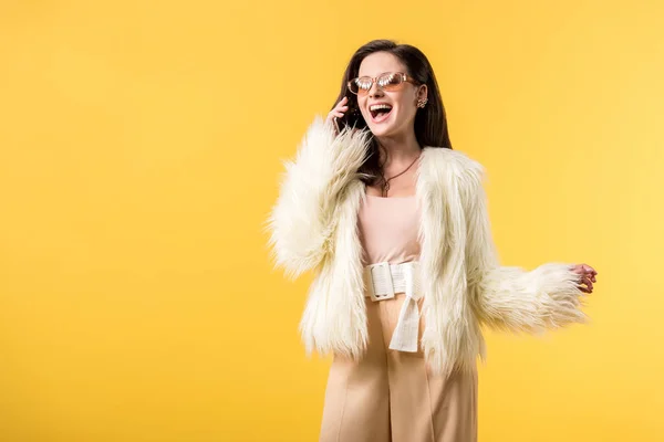 Happy party girl in faux fur jacket and sunglasses talking on smartphone isolated on yellow — Stock Photo