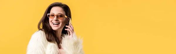 Happy party girl en fausse fourrure et lunettes de soleil parlant sur smartphone isolé sur jaune, plan panoramique — Photo de stock