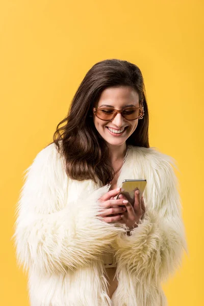 Happy party girl in faux fur jacket and sunglasses using smartphone isolated on yellow — Stock Photo