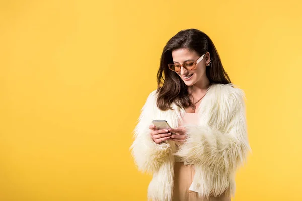 Chica de fiesta feliz en chaqueta de piel sintética y gafas de sol usando smartphone aislado en amarillo - foto de stock