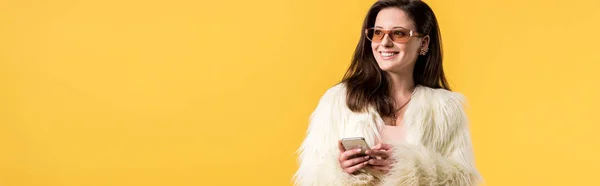 Chica fiesta feliz en chaqueta de piel sintética y gafas de sol celebración de teléfono inteligente aislado en amarillo, tiro panorámico — Stock Photo