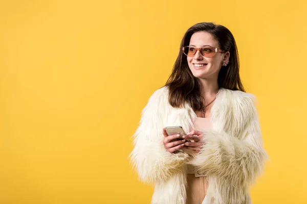 Chica de fiesta feliz en chaqueta de piel sintética y gafas de sol con teléfono inteligente aislado en amarillo - foto de stock