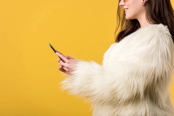 Vista recortada de chica de fiesta feliz en chaqueta de piel sintética y gafas de sol con teléfono inteligente aislado en amarillo - foto de stock