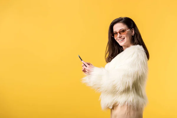 Chica de fiesta feliz en chaqueta de piel sintética y gafas de sol con teléfono inteligente aislado en amarillo - foto de stock