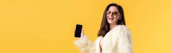 Colpo panoramico di festa felice ragazza in giacca di pelliccia ecologica e occhiali da sole in possesso di smartphone con schermo bianco isolato su giallo — Foto stock