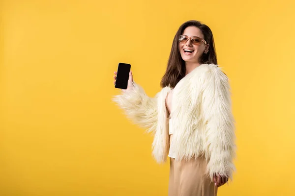 Happy party girl veste en fausse fourrure et lunettes de soleil tenant smartphone avec écran blanc isolé sur jaune — Photo de stock