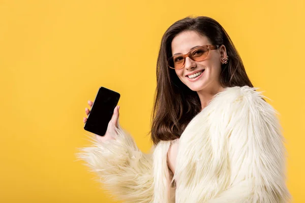 Happy party girl in faux fur jacket and sunglasses holding smartphone with blank screen isolated on yellow — Stock Photo