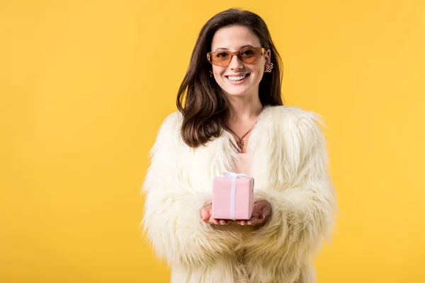 Chica de fiesta feliz en chaqueta de piel sintética y gafas de sol con caja de regalo aislado en amarillo - foto de stock