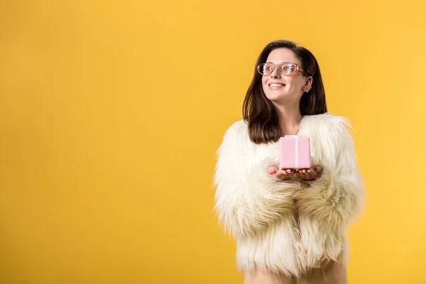 Menina festa feliz em casaco de pele falso e óculos de sol segurando caixa de presente e olhando para longe isolado no amarelo — Fotografia de Stock