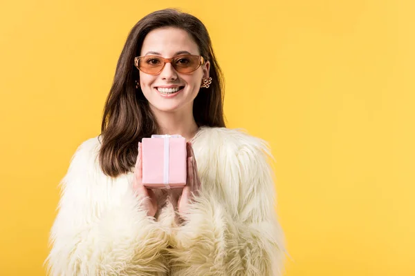 Felice festa ragazza in giacca di pelliccia sintetica e occhiali da sole in possesso di scatola regalo isolato su giallo — Foto stock