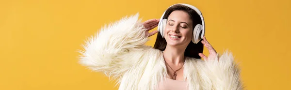 Panoramic shot of smiling girl with closed eyes in faux fur jacket listening music in headphones isolated on yellow — Stock Photo