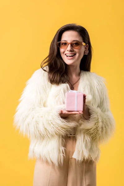 Felice festa ragazza in giacca di pelliccia sintetica e occhiali da sole in possesso di scatola regalo isolato su giallo — Foto stock