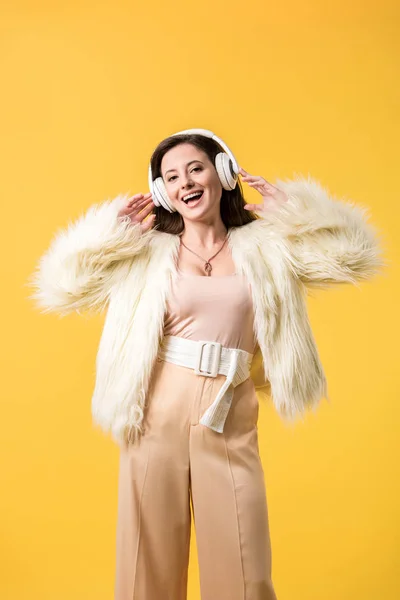 Улыбающаяся девушка в искусственном меховом пиджаке слушает музыку в наушниках, изолированных на желтом — стоковое фото