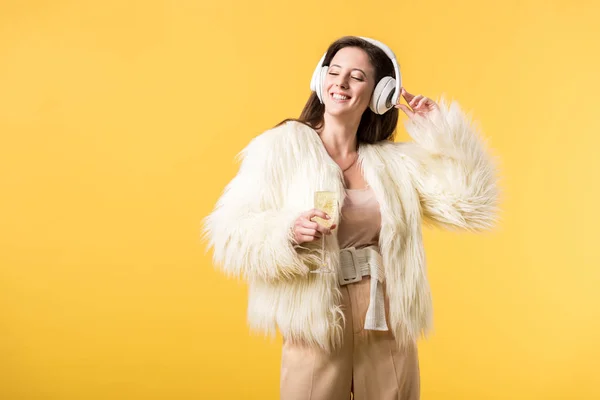 Улыбающаяся девушка в искусственном меховом пиджаке слушает музыку в наушниках и держит бокал шампанского изолированным на желтом — стоковое фото